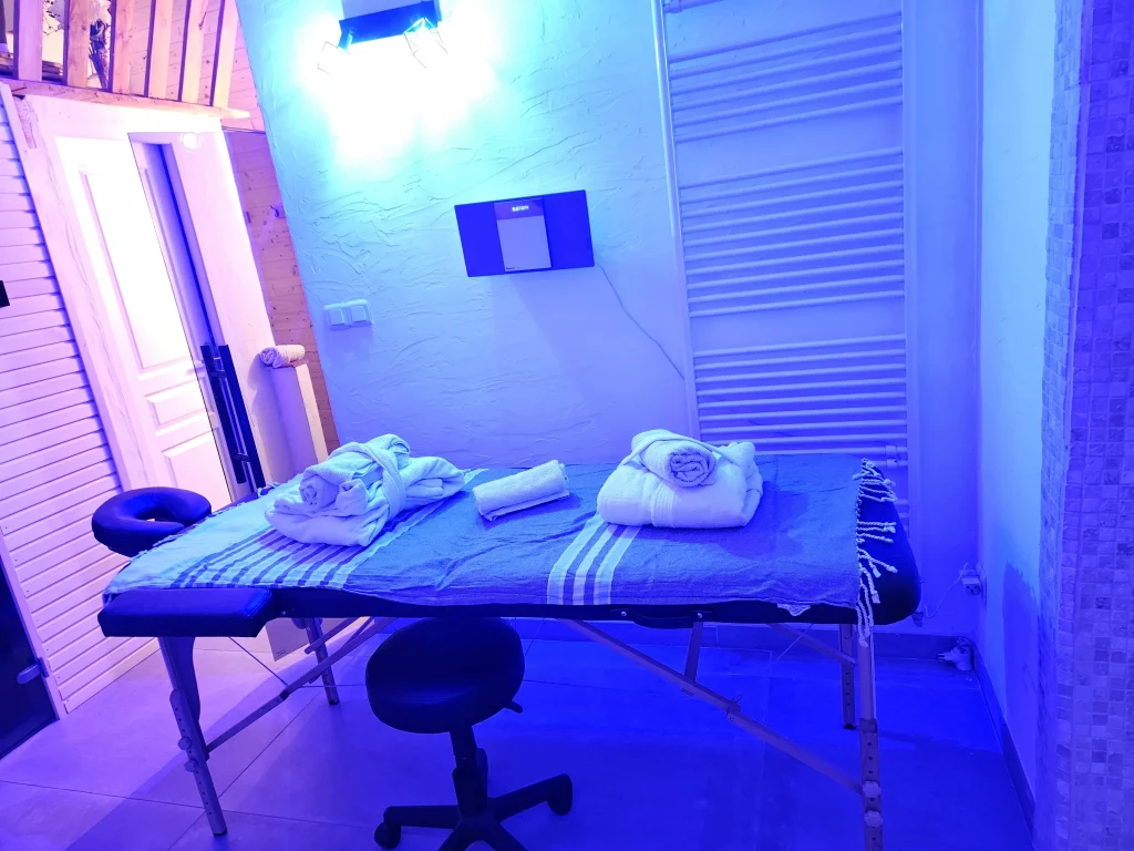 Salle de massage avec un lit simple, serviettes blanches et lumière bleue pour une atmosphère relaxante.